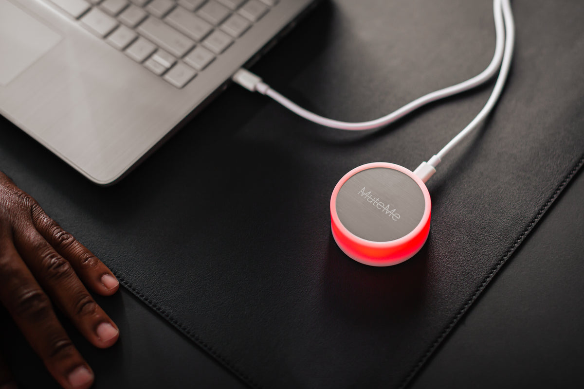 MuteMe Original glowing red on a dark desk beside a laptop and a resting hand.