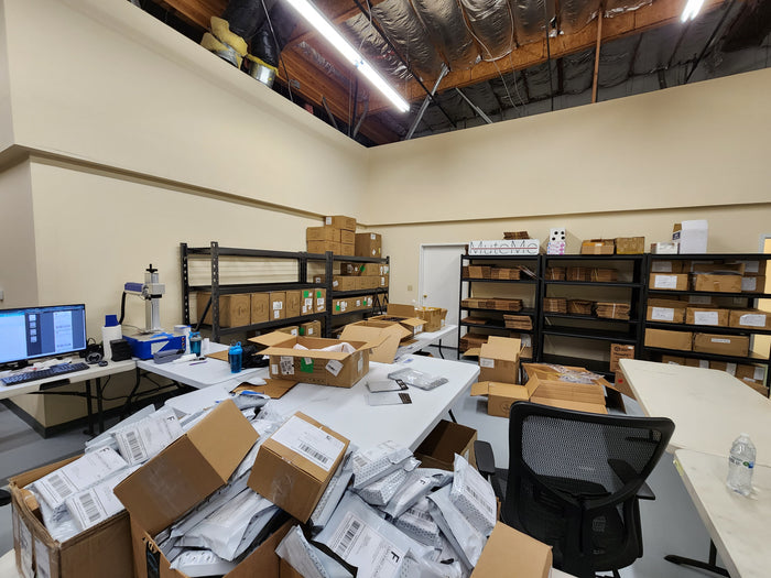 A bunch of packages that need to be mailed out, with a laser engraver in the background and some packaging materials on shelves in the background.